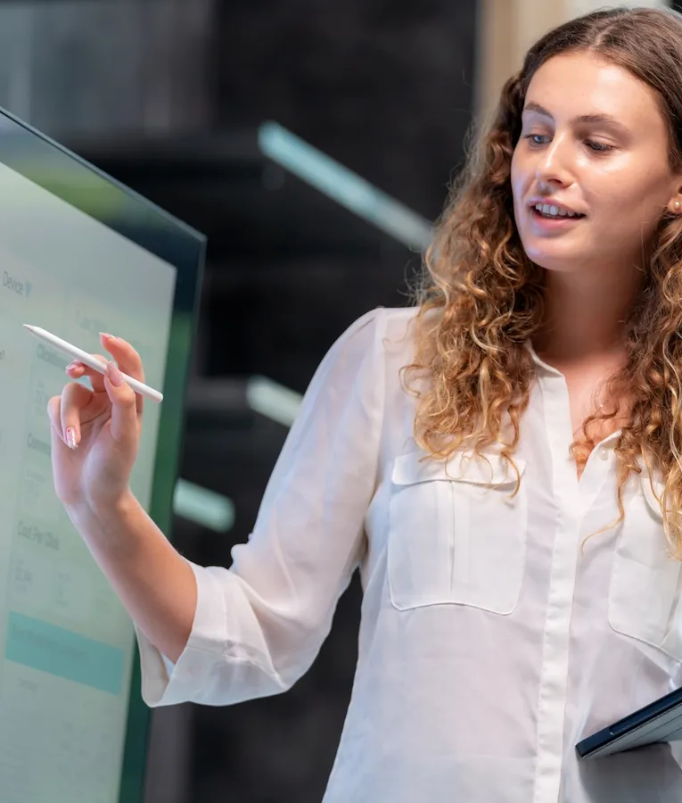 A professional woman with curly hair in a white blouse presents data analytics on a large touchscreen, using a stylus while holding a tablet in a modern office.