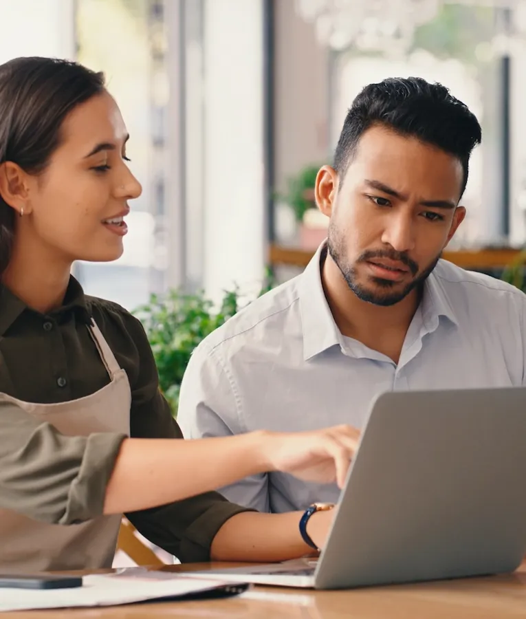 En cafémedarbejder i forklæde og en ejer gennemgår økonomiske dokumenter sammen på en bærbar computer i en moderne café.