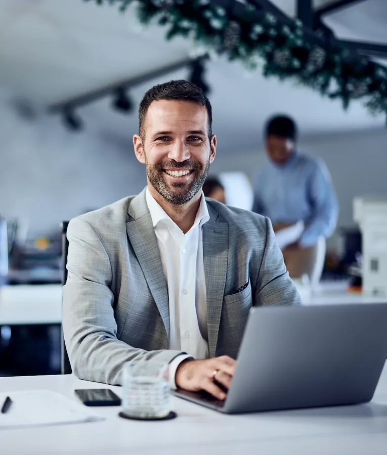 Smiling businessman working on a laptop in a modern office, with colleagues collaborating in the background. Represents workplace productivity and workforce optimization.