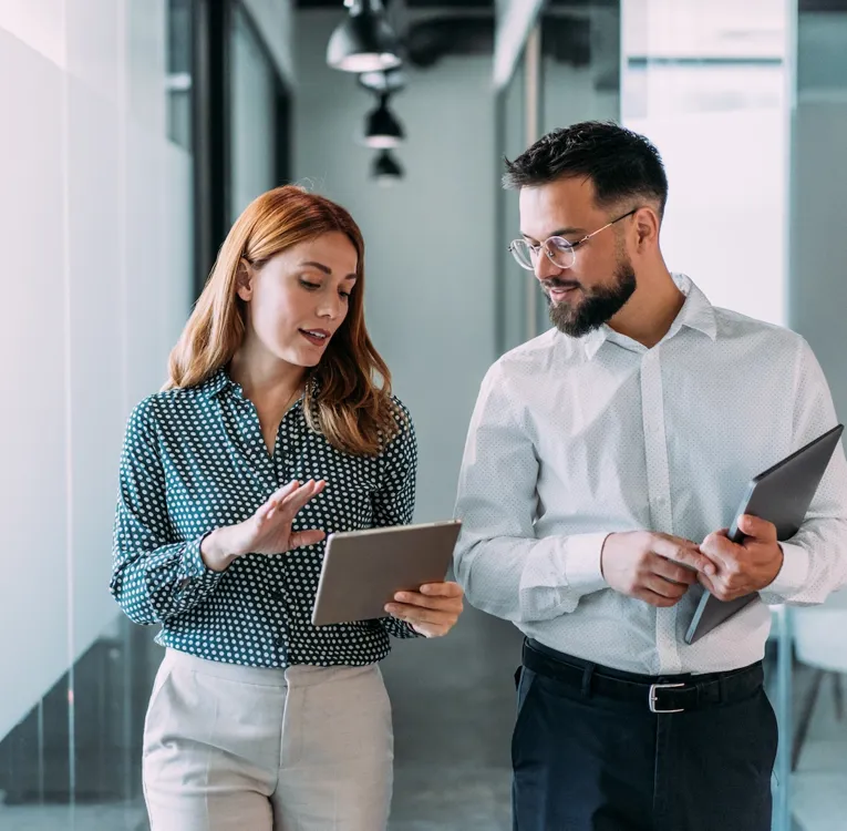 Two business professionals walking through a modern office, discussing data on tablets. Represents mobile workforce collaboration and digital efficiency.