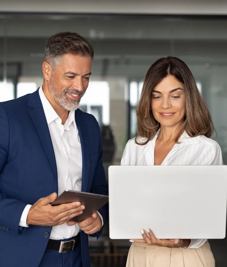 Two smiling business professionals collaborating in a modern office, reviewing data on a laptop and tablet. Represents teamwork, leadership, and digital workforce solutions.