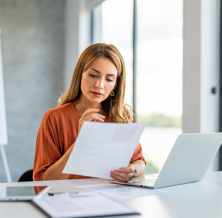 A woman with long light brown hair, wearing an orange blouse, is reviewing a document while working on a laptop in a bright, modern office. A tablet and clipboard are placed on the desk, and a whiteboard with graphs is visible in the background. Ideal for themes like data analysis, business strategy, and productivity.