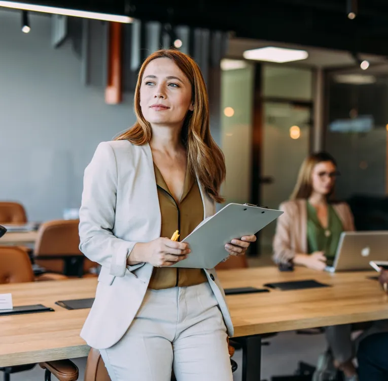 A confident businesswoman in a beige suit holds a clipboard while overseeing a workforce strategy meeting in a modern, well-lit office.