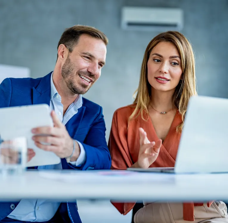 A man in a blue suit and a woman in a rust-colored blouse collaborate in a modern office, analyzing workforce data on a laptop and tablet.