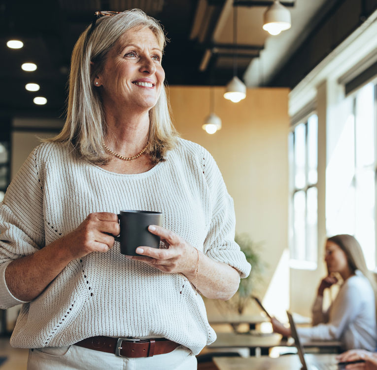 A smiling senior woman with gray hair, wearing a cozy sweater, holds a coffee cup while standing in a modern coworking space. In the background, professionals are working on laptops, creating a dynamic and collaborative atmosphere.