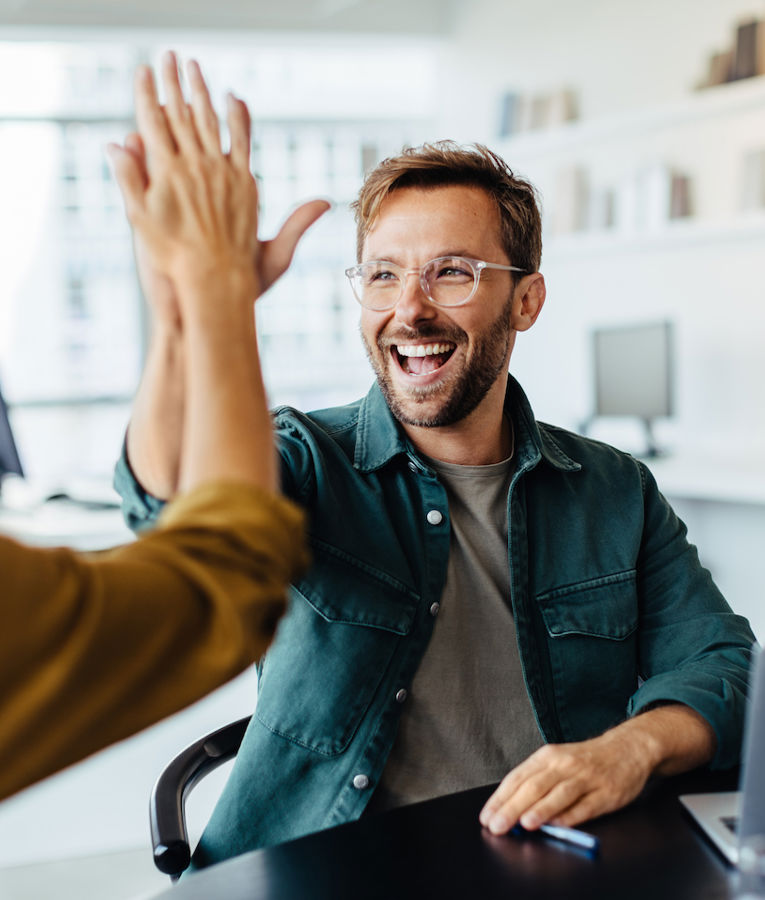 A cheerful team in a modern office celebrates a success as a man and a woman exchange a high-five. Another colleague smiles while working on a laptop, creating a positive and collaborative work atmosphere.
