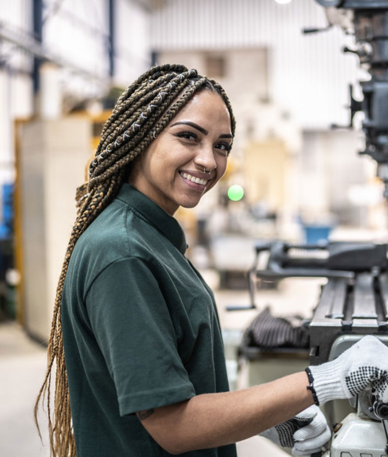 A smiling female machinist in a dark green uniform and gloves operating a milling machine in a manufacturing workshop.
