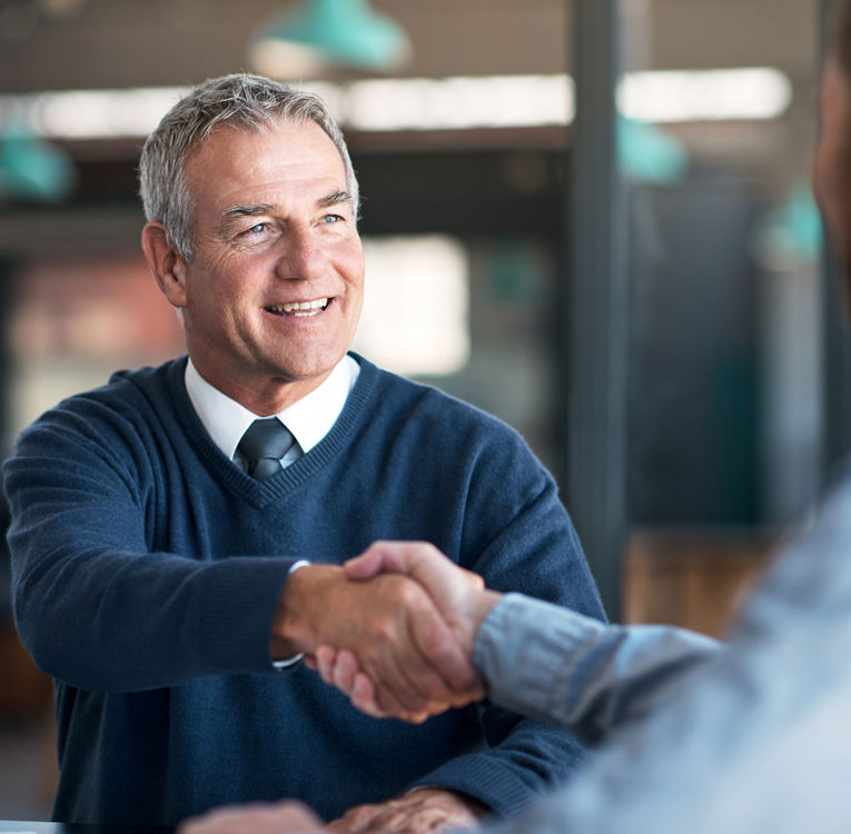 A smiling senior businessman in a navy sweater and tie shakes hands with a colleague in a modern office or café setting. The handshake signifies a successful deal or agreement, reflecting professionalism and trust.