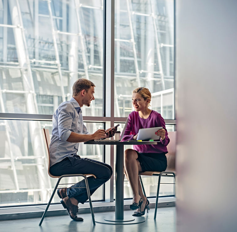 A man and a woman engaged in a business discussion at a small round table in a modern office with large glass windows. The man is holding a smartphone, while the woman, dressed in a purple blouse, holds a tablet and smiles.