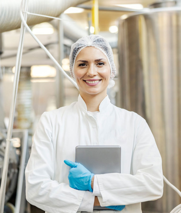 A smiling female quality control specialist in a white lab coat, hairnet, and gloves holding a tablet in a modern food production facility.