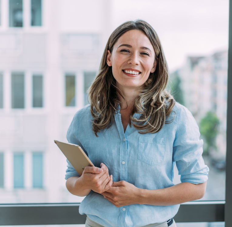 A cheerful young woman with long brown hair, wearing a light blue blouse, stands in a modern office holding a tablet. She smiles confidently, with large windows and an urban cityscape in the background.