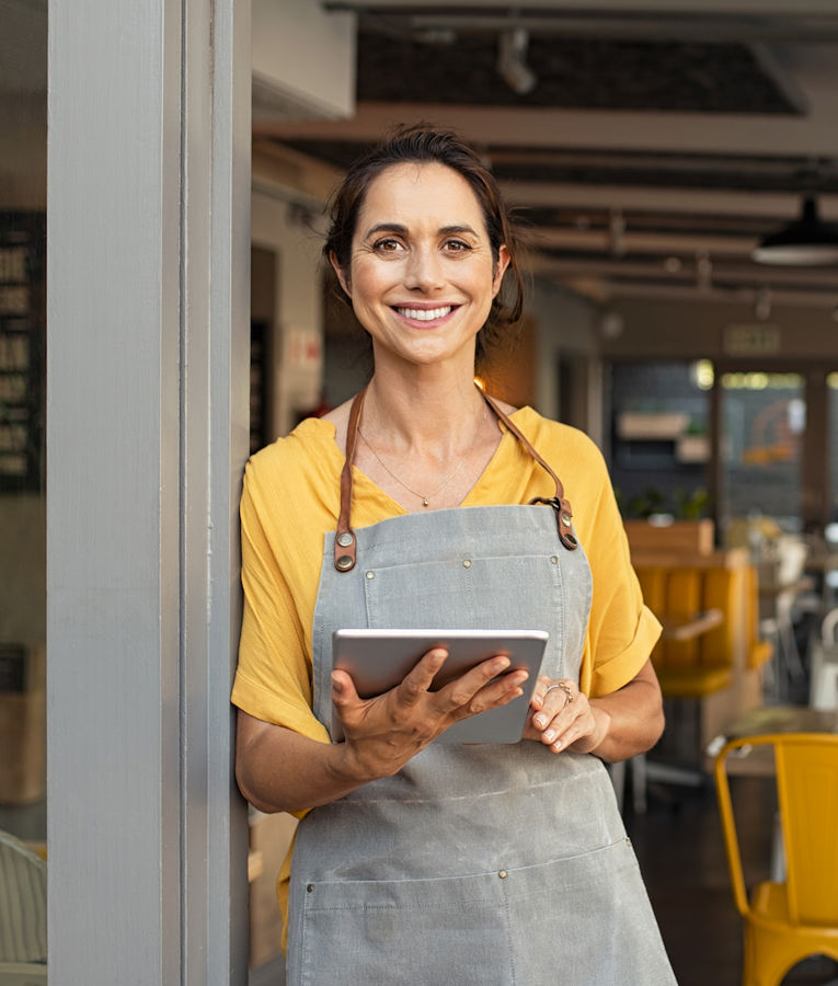 A cheerful female restaurant owner or manager wearing a yellow blouse and a gray apron stands at the entrance of a busy café, holding a tablet. She looks confident and welcoming, managing daily operations and customer service.