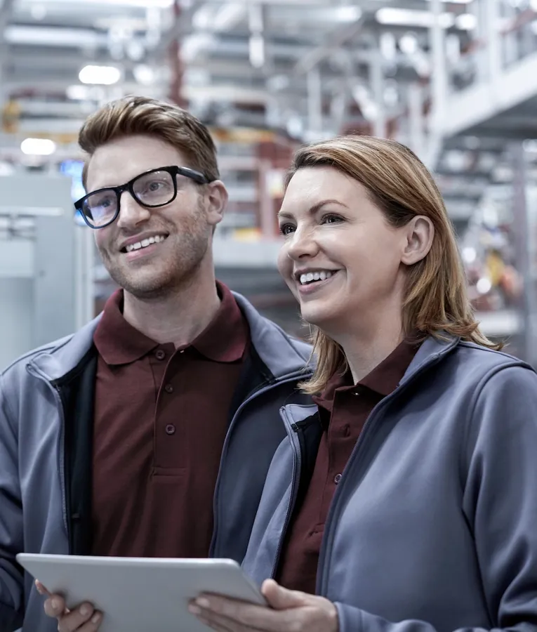 Two smiling factory workers using a tablet and monitoring a control panel in an advanced manufacturing facility. Represents workforce automation and industrial efficiency.