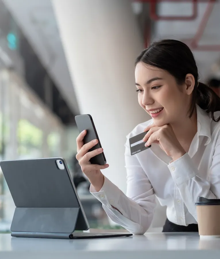Smiling businesswoman using a smartphone and credit card for an online transaction, with a tablet and coffee on the table, representing secure digital payments.