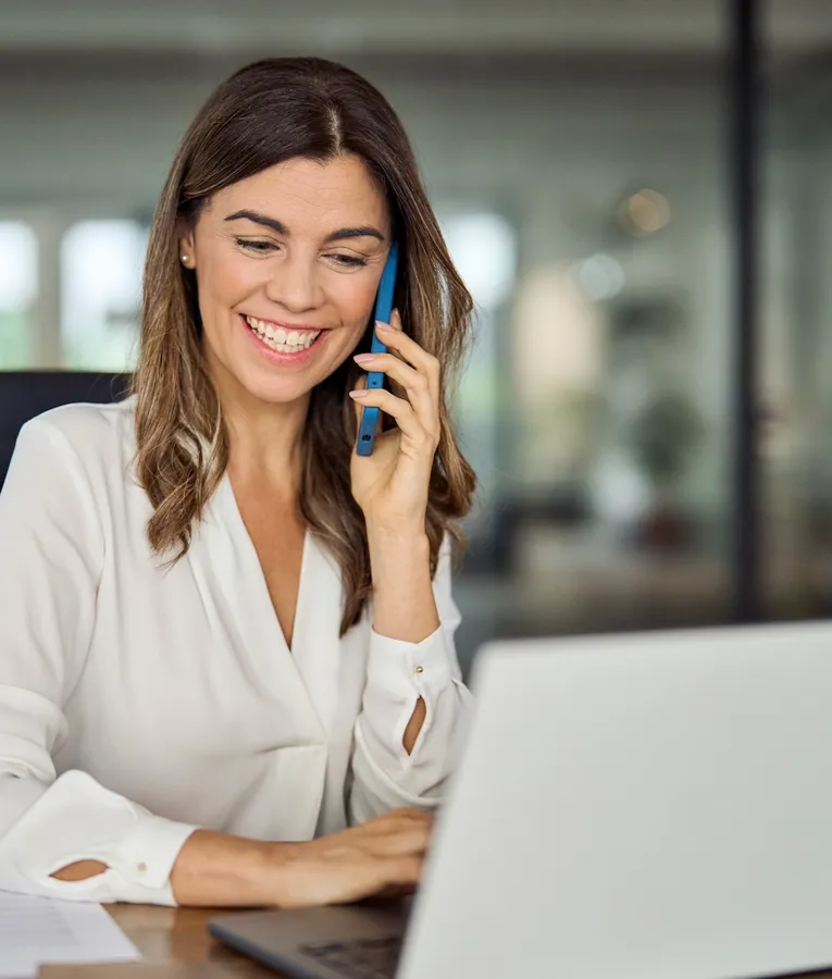 A cheerful businesswoman in a white blouse sits at her desk, talking on the phone while using her laptop. The image conveys productivity, communication, and efficient workplace management.