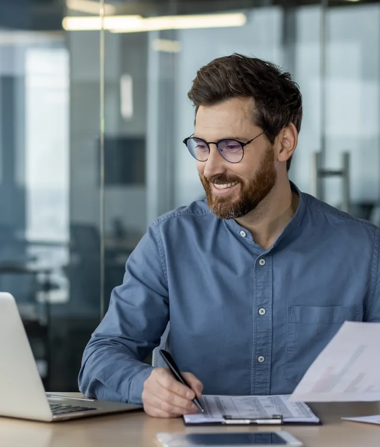 Smiling businessman analyzing charts and reports at his desk, using a laptop for workforce planning and data-driven decision-making. Represents efficient HR and business management.