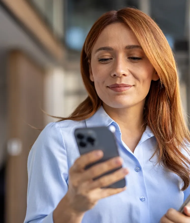 Confident businesswoman using a smartphone while holding a tablet, managing tasks remotely. Represents mobile workforce management and digital efficiency.