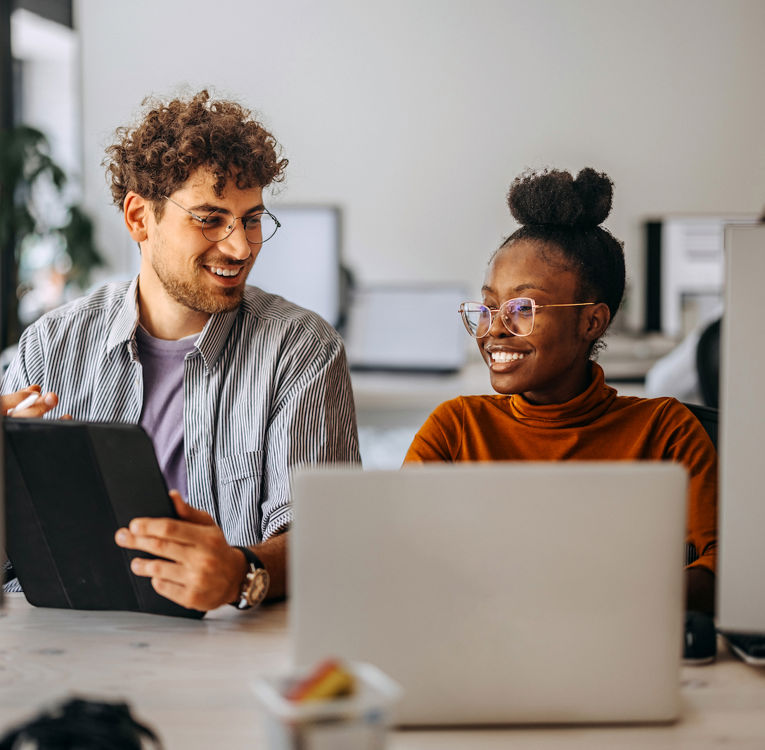 A smiling man holding a tablet and a woman working on a laptop are having a friendly discussion in a modern office setting. Both are wearing glasses and appear engaged in a collaborative project. Perfect for themes related to teamwork, productivity, and office collaboration.