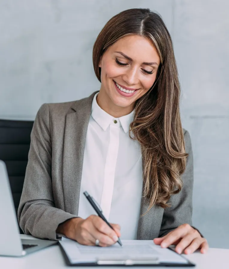 A smiling businesswoman in a grey blazer signing documents at her desk with a laptop beside her.