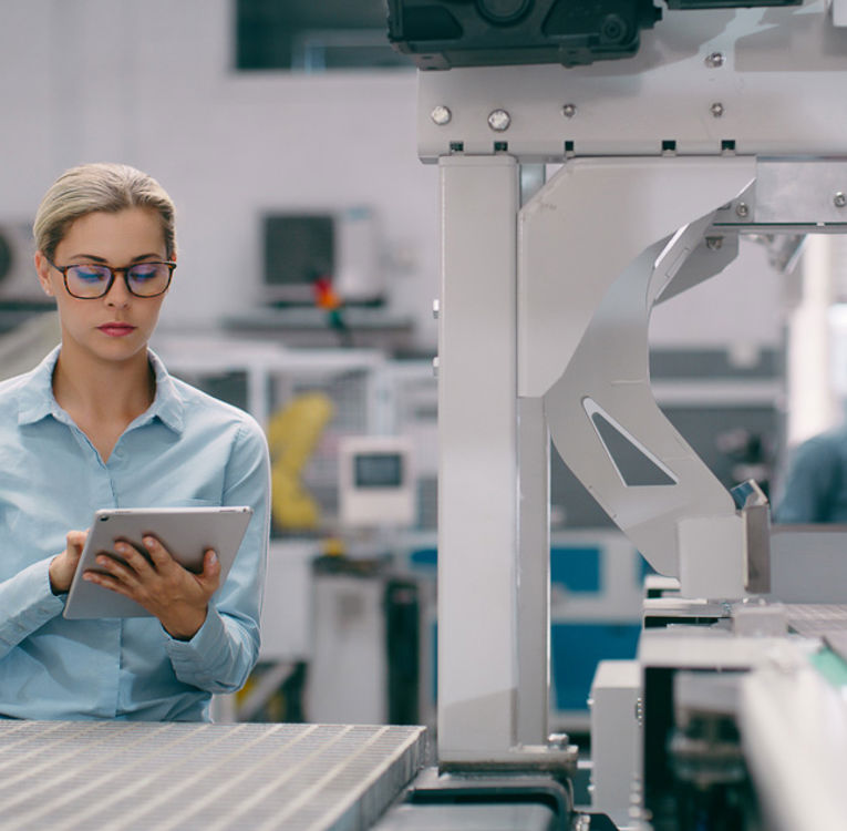 A focused female engineer in a light blue shirt using a tablet to monitor a production line in a modern industrial facility.