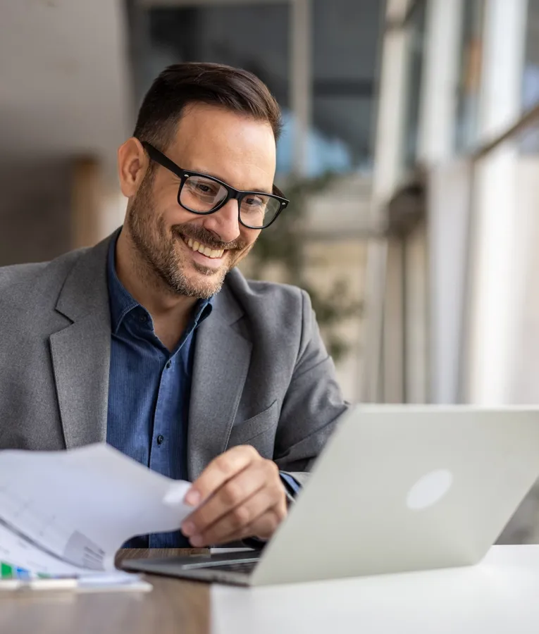 A smiling business professional in a grey blazer and glasses reviews workforce analytics on a laptop while holding financial reports in a modern office setting.