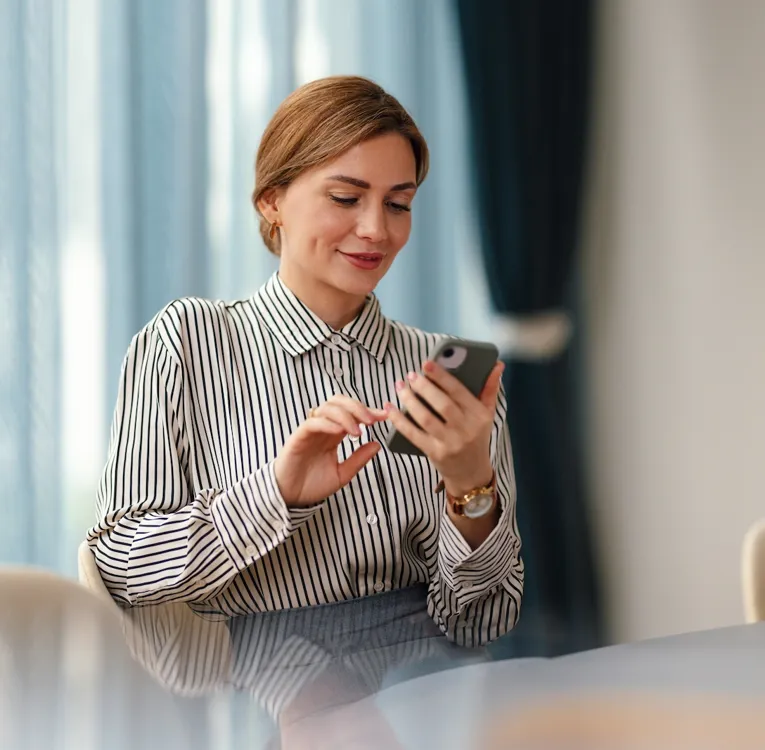 A professional woman in a striped blouse sits at a conference table, smiling while using her smartphone in a modern office with blue curtains.