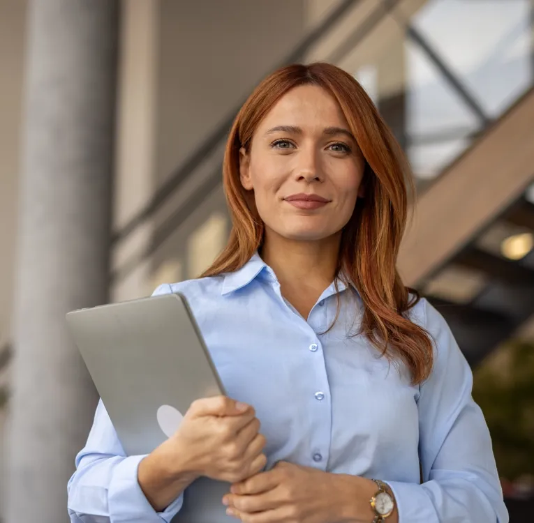 A confident businesswoman with red hair, wearing a light blue blouse, holds a laptop while standing in a modern office. The image represents leadership, technology, and efficient workforce management.