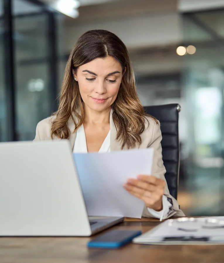 Focused businesswoman reviewing documents at her desk with a laptop, representing efficient workforce management and business administration.