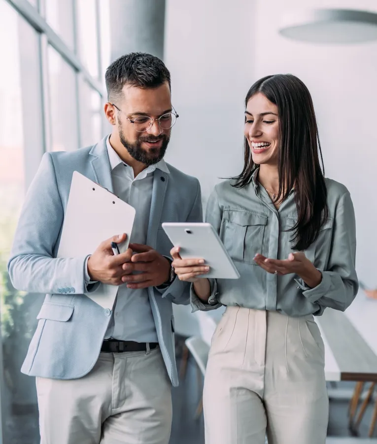 A smiling businesswoman with a tablet discusses workforce management with a male colleague holding a clipboard in a bright, modern office.