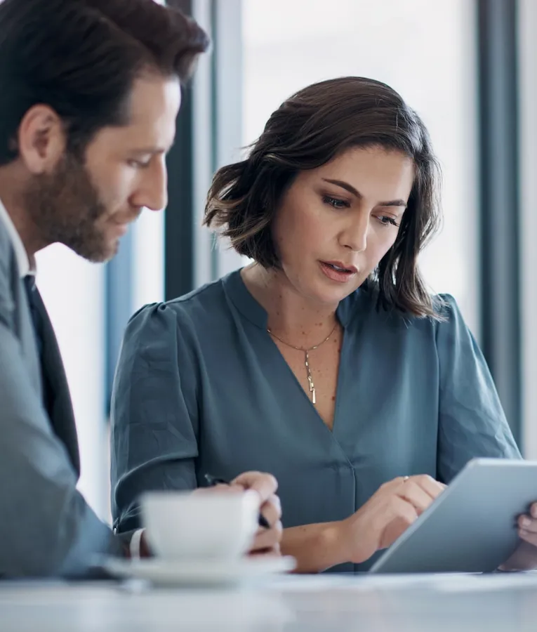A professional woman in a blue blouse uses a tablet to discuss work with a male colleague in a grey suit over coffee in a modern office setting.