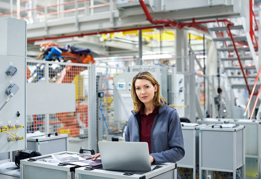 A confident female engineer working on a laptop in a modern industrial facility, monitoring automated production systems.