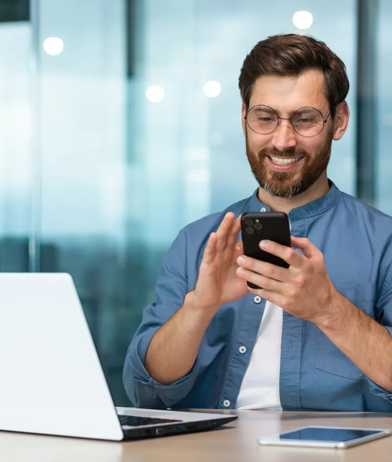 Smilende forretningsmand bruger en smartphone ved sit skrivebord med en bærbar og tablet, der viser mobil arbejdsstyring på et moderne kontor.