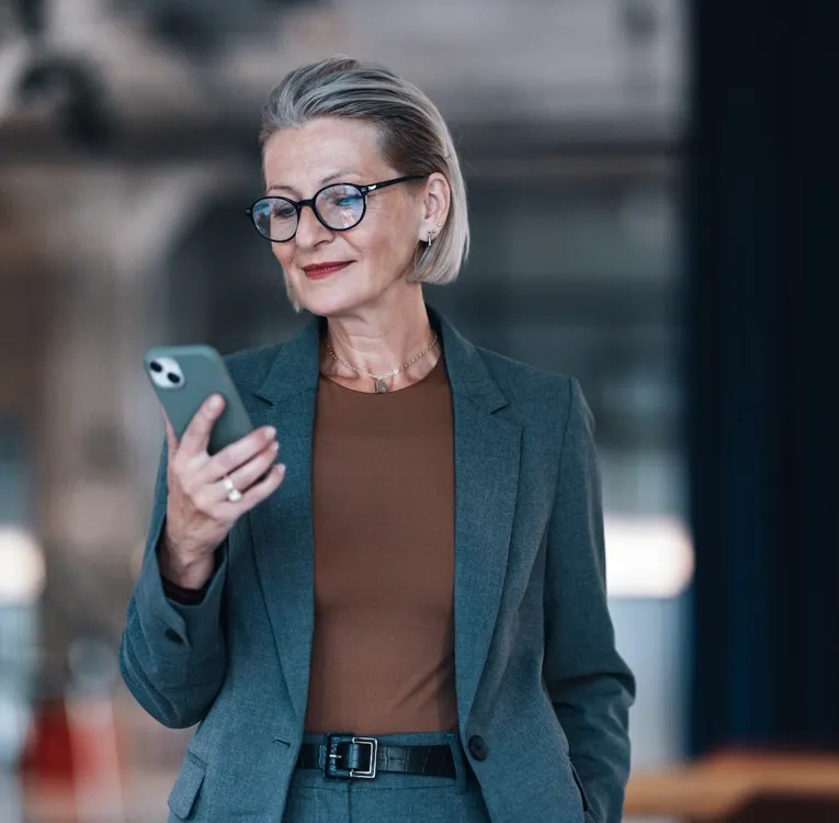 Selvsikker senior forretningskvinde bruger en smartphone på et moderne kontor til at styre opgaver og arbejdsstyrkeoperationer eksternt. Symboliserer lederskab og mobil produktivitet.