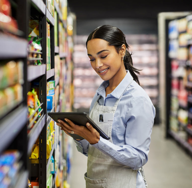 A smiling supermarket employee in a blue shirt and apron uses a digital tablet to check inventory while working in the grocery store aisle.