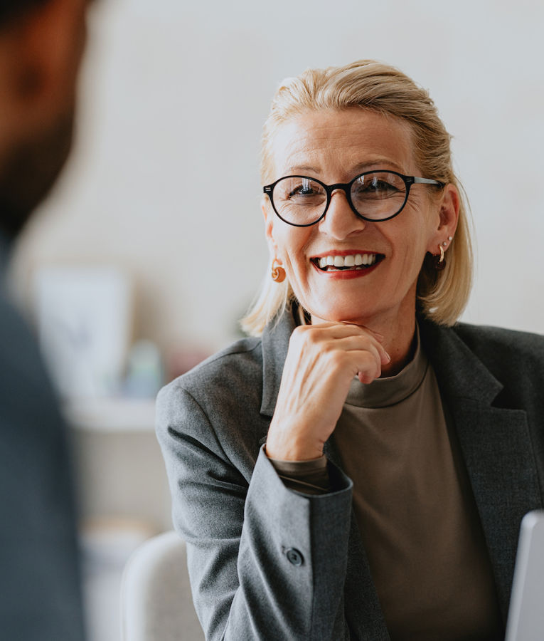A smiling woman with short blonde hair and round glasses, dressed in a gray blazer, is engaged in a conversation in a professional setting. She appears confident and approachable, with a laptop in front of her. Perfect for themes like leadership, business consultation, and executive meetings.