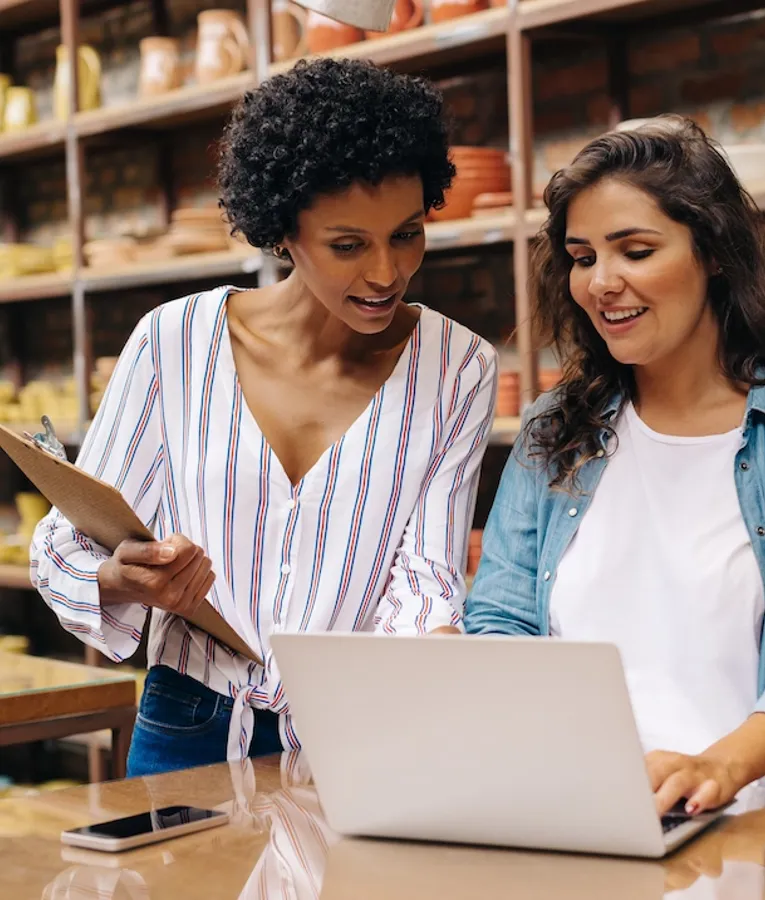 To smilende butiksmedarbejdere arbejder sammen ved en butikdisk og bruger en bærbar computer til at administrere lager og salg omgivet af keramikprodukter.