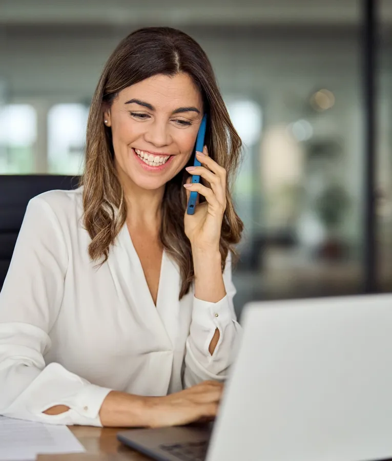 Smiling businesswoman talking on the phone while working on a laptop in a modern office, representing efficient remote workforce communication.