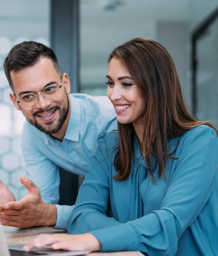 Two smiling business professionals collaborating on a laptop in a modern office, discussing strategies and workforce management. Represents teamwork and digital productivity.